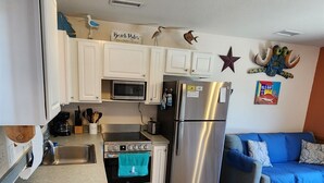 A compact kitchen with white cabinets, a stainless steel refrigerator, a microwave, and a stove. There is a blue couch in the adjacent living area. - Put your Anchor down with Nautical Nine! Beachfront, Pet Friendly Cabana! (HATTERAS)