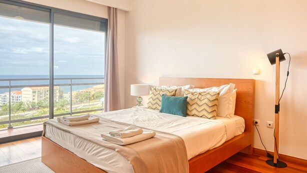A hotel room with a large bed, a nightstand, a lamp, and a view of the sea. - Levada dos Piornais, a Home in Madeira (Funchal)