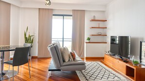 A modern living room with a grey sofa, a dining table with chairs, a TV, and wooden shelves. - Levada dos Piornais, a Home in Madeira (Funchal)