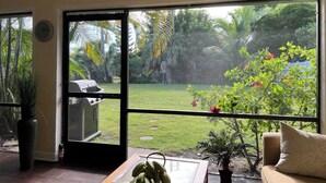 A room with a large sliding glass door leading to a garden. Inside, there is a wooden table with a plant, a couch, and a small potted plant.