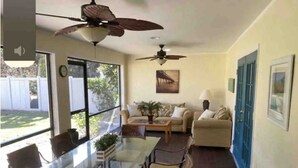 A spacious room with a glass dining table, comfortable seating, ceiling fans, and large windows offering a view of the outdoors.