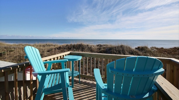 Um deck de madeira com cadeiras Adirondack azuis e uma pequena mesa, com vista para uma praia com dunas e o oceano.