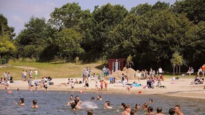 Ein überfüllter Strand mit badenden und sonnenden Menschen, eine kleine Bauskonstruktion und ein bewaldeter Hügel im Hintergrund.
