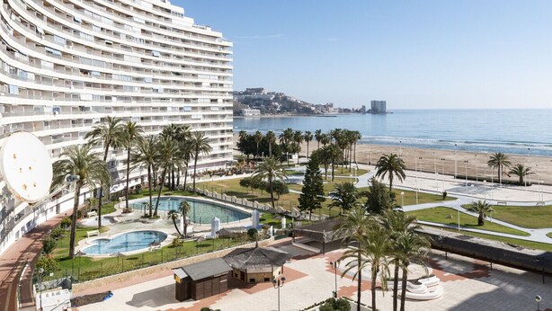 Ein Hotel mit geschwungener Fassade, einem Pool, Palmen und Blick auf den Strand.