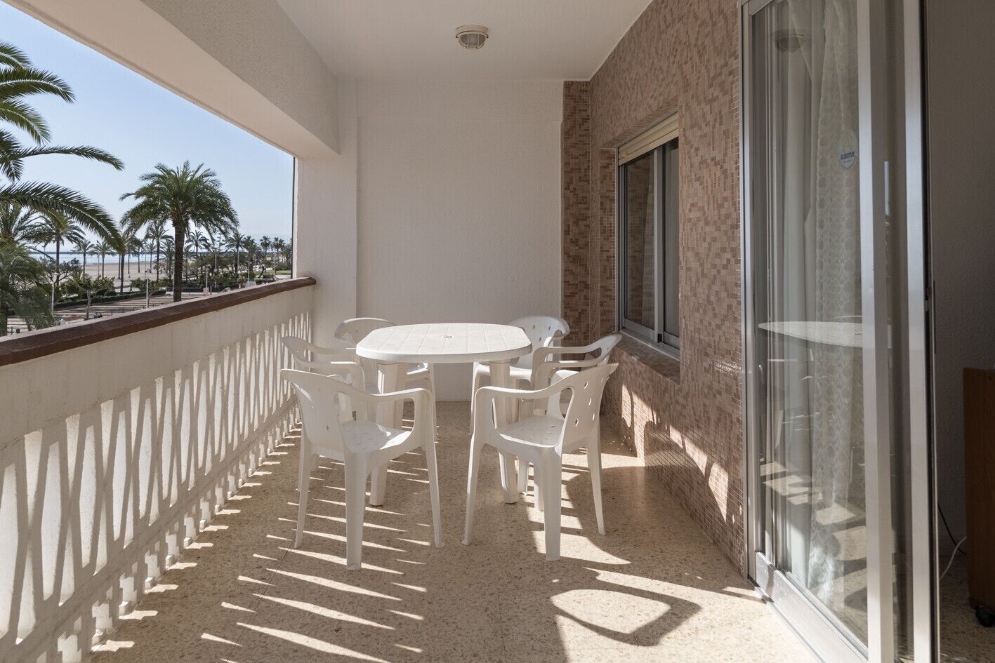 A balcony with a round table and chairs, a view of palm trees, and a tiled floor.