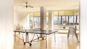 Ein geräumiger Raum mit Tischtennisplatte, weißen Stühlen, Deckenventilator, großen Fenstern mit Meerblick und einem Balkon mit weißen Stühlen.