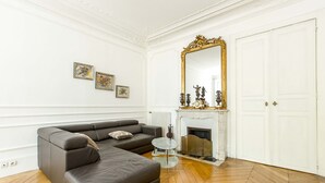 A living room with a black leather sofa, a white fireplace, a large mirror, and a herringbone wooden floor. - By Boulevard Saint Germain, by Veeve (Paris)