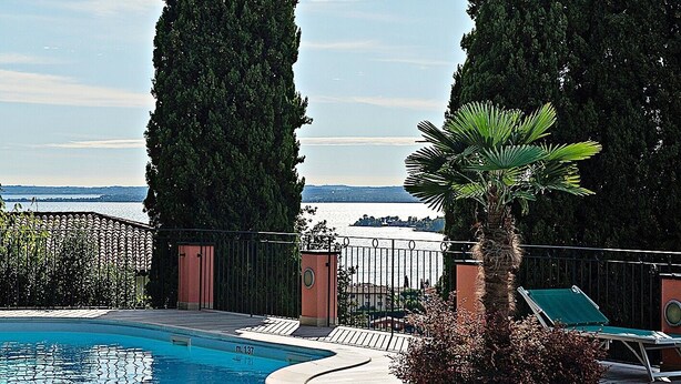 A swimming pool with a view of the water, surrounded by tall trees and a fence. - Beautiful View in a Shabby Chic Atmosphere (Padenghe Sul Garda)