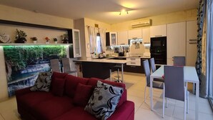 A modern living room with a red sofa, a kitchen area with white cabinets, and a dining space with a glass table. - IL-BEJTA  PENTHOUSE. (Il-Qala)