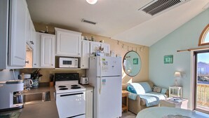 A compact kitchen with white cabinets, a microwave, a refrigerator, and a stove. There is a dining area with a table and chairs, and a window with a view of the outside. - Escape to the beach with Cabana By The Sea! (HATTERAS)