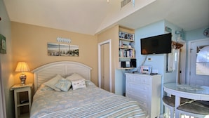 A bedroom with a bed, a bedside table with a lamp, a TV mounted on the wall, a bookshelf, and a small table with chairs. - Escape to the beach with Cabana By The Sea! (HATTERAS)