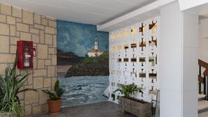 Un hall d’entrée orné d’une fresque représentant un paysage côtier, avec un extincteur, des plantes en pot et un mur décoratif blanc.