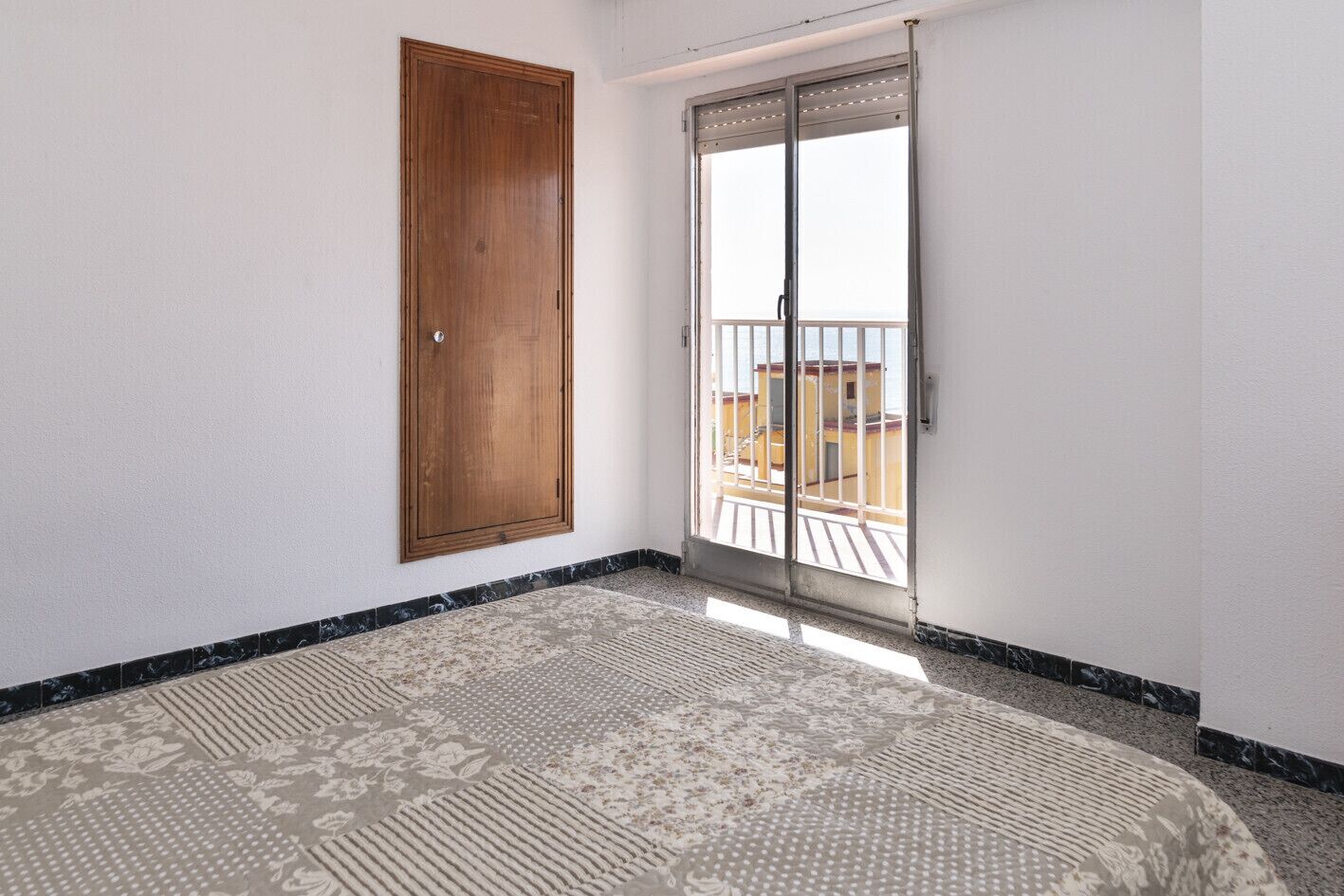 A room with a wooden door, a sliding glass door leading to a balcony, and a patterned floor.
