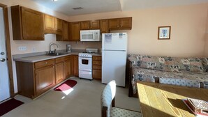 A compact kitchen with wooden cabinets, a microwave, a refrigerator, and a stove. There is a small dining area with a table and a patterned sofa. - A Dune Away is the place to stay! (HATTERAS)