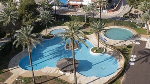 Une grande piscine extérieure, bordée de palmiers, entourée d’une allée pavée et avec un bâtiment en arrière-plan.