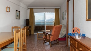 Habitación con una mesa de comedor, sillas, un sofá, un televisor y un balcón con vistas al mar y a las montañas.
