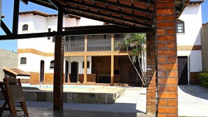 A covered outdoor area with a pool, brick pillars, and a white building with balconies in the background. - Beautiful 5 Bedrooms (4 Suites) Air conditioning, swimming pool, barbecue - Saquarema (Saquarema)
