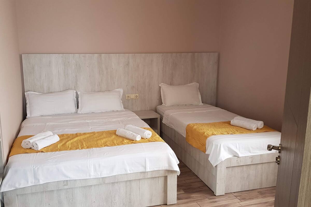 A hotel room with two single beds, each with white bedding and a yellow throw blanket. There is a wooden headboard, a small bedside table, and a mirror on the left wall.