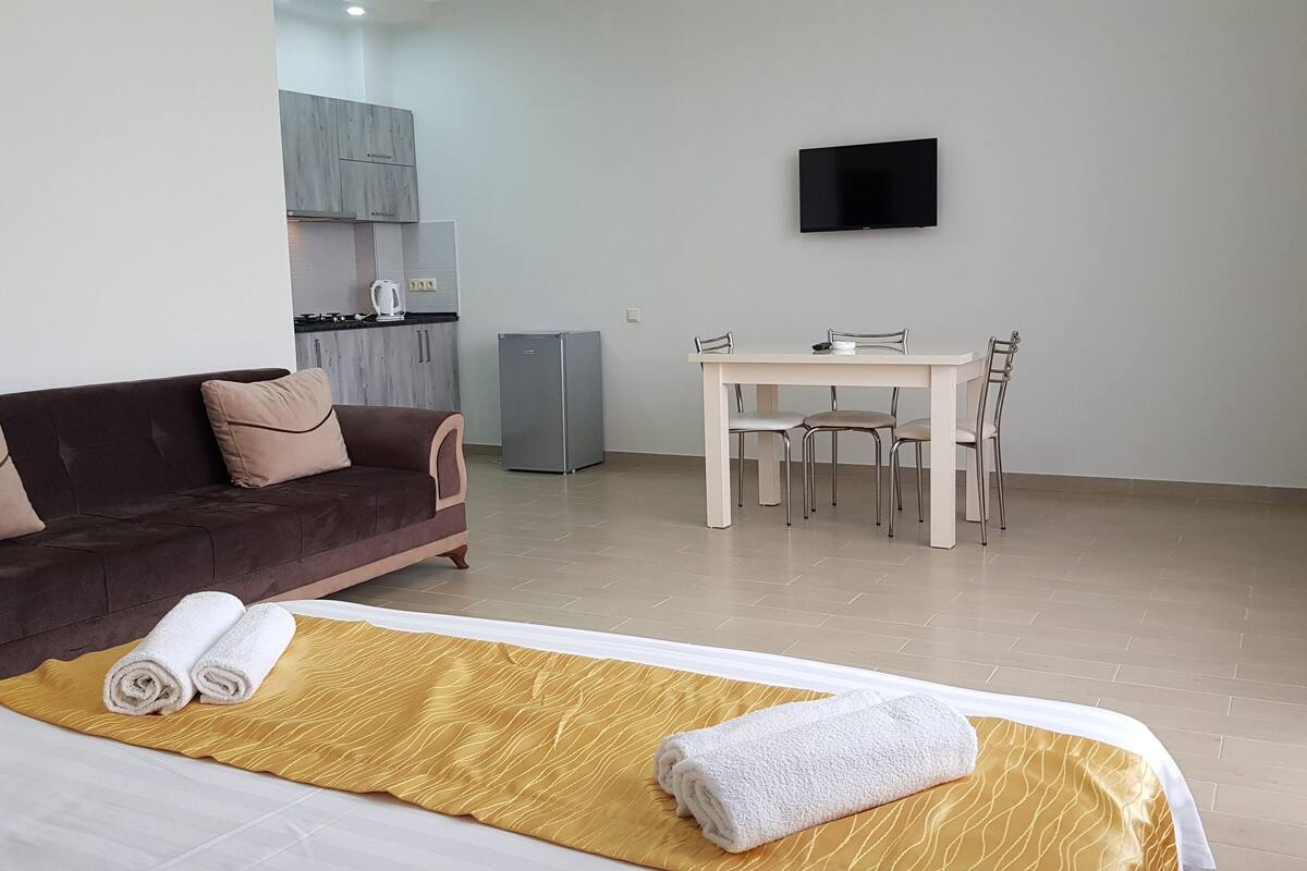A hotel room with a bed, a sofa, a dining table with chairs, a TV on the wall, and a kitchen area in the background.