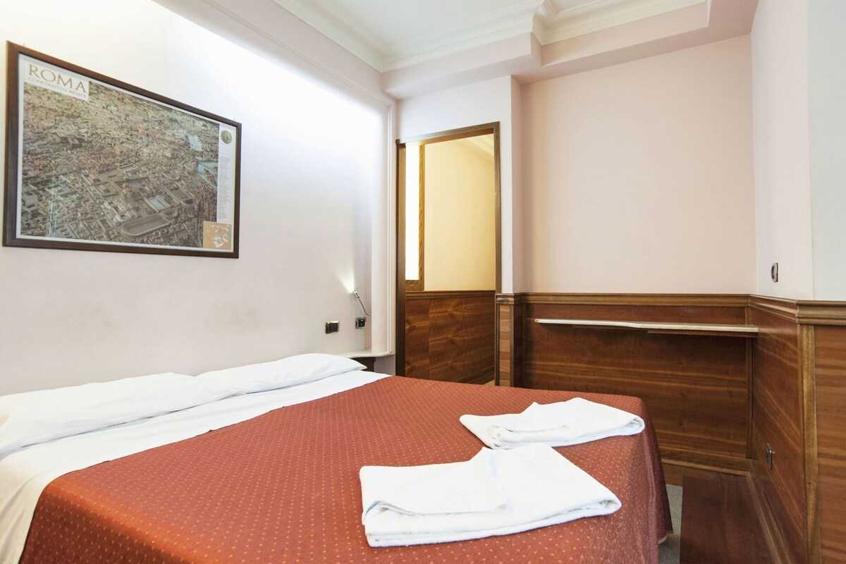 A hotel room with a bed, a wooden dresser, a mirror, and a framed map of Rome on the wall.