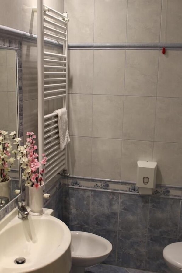 Un baño con un lavamanos blanco, dos inodoros, un perchero para toallas y un dispensador de jabón.