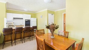 Een compacte keuken met witte kastjes, een magnetron, een oven en een koelkast. Er is een houten eettafel met zes stoelen en een vaas met gedroogde bloemen.