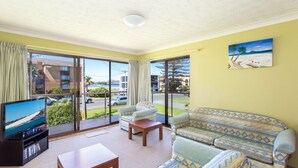 Een woonkamer met een televisie, bank en uitzicht op een strand door grote ramen.