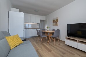A modern living space with a grey sofa, a yellow pillow, a dining area with a table and chairs, a kitchen with white cabinets, and a flat-screen TV. - Apartment Tara Apt 3 (Medulin)
