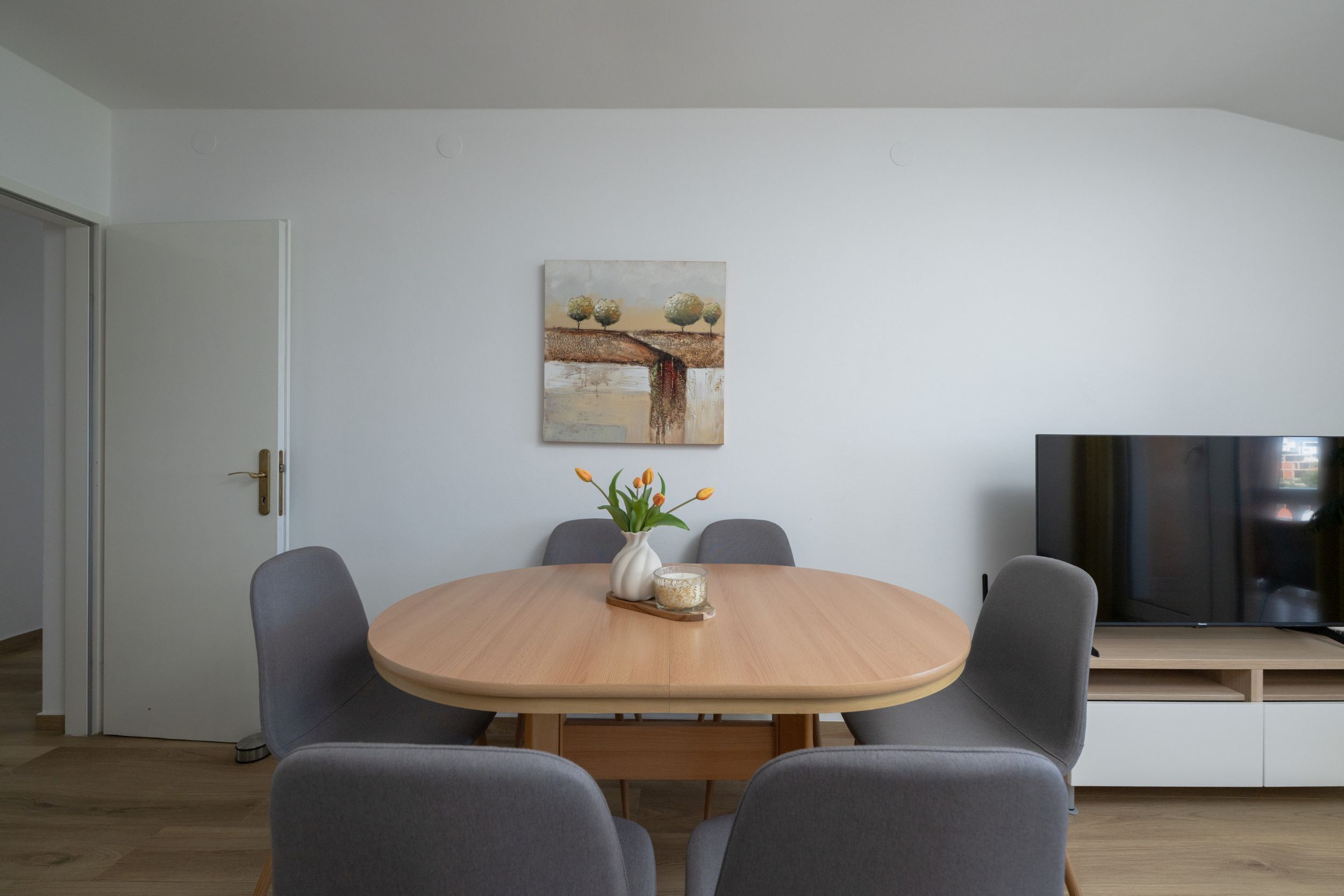 A dining area with a round table, grey chairs, a vase with flowers, and a painting on the wall.