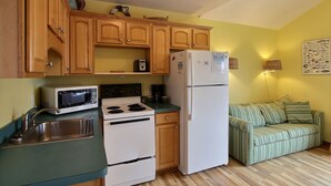 Una cucina compatta con mobili in legno, un forno a microonde, fornelli e un frigorifero. Nell'area soggiorno adiacente, un divano verde.