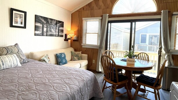 A bedroom with a bed, a sofa, a dining table with chairs, a window with curtains, and a wall with framed pictures.