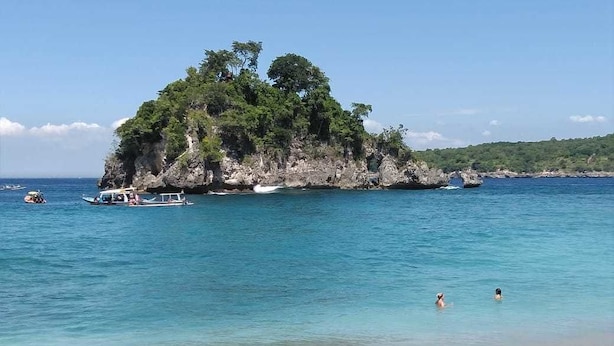 A beach with clear turquoise water, a small island with dense greenery, and a few boats. - Arie Guest House Nusa Penida (Penida Island)