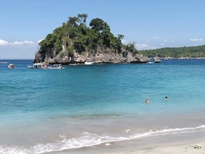 A beach with clear turquoise water, a small island with dense greenery, and a few boats.