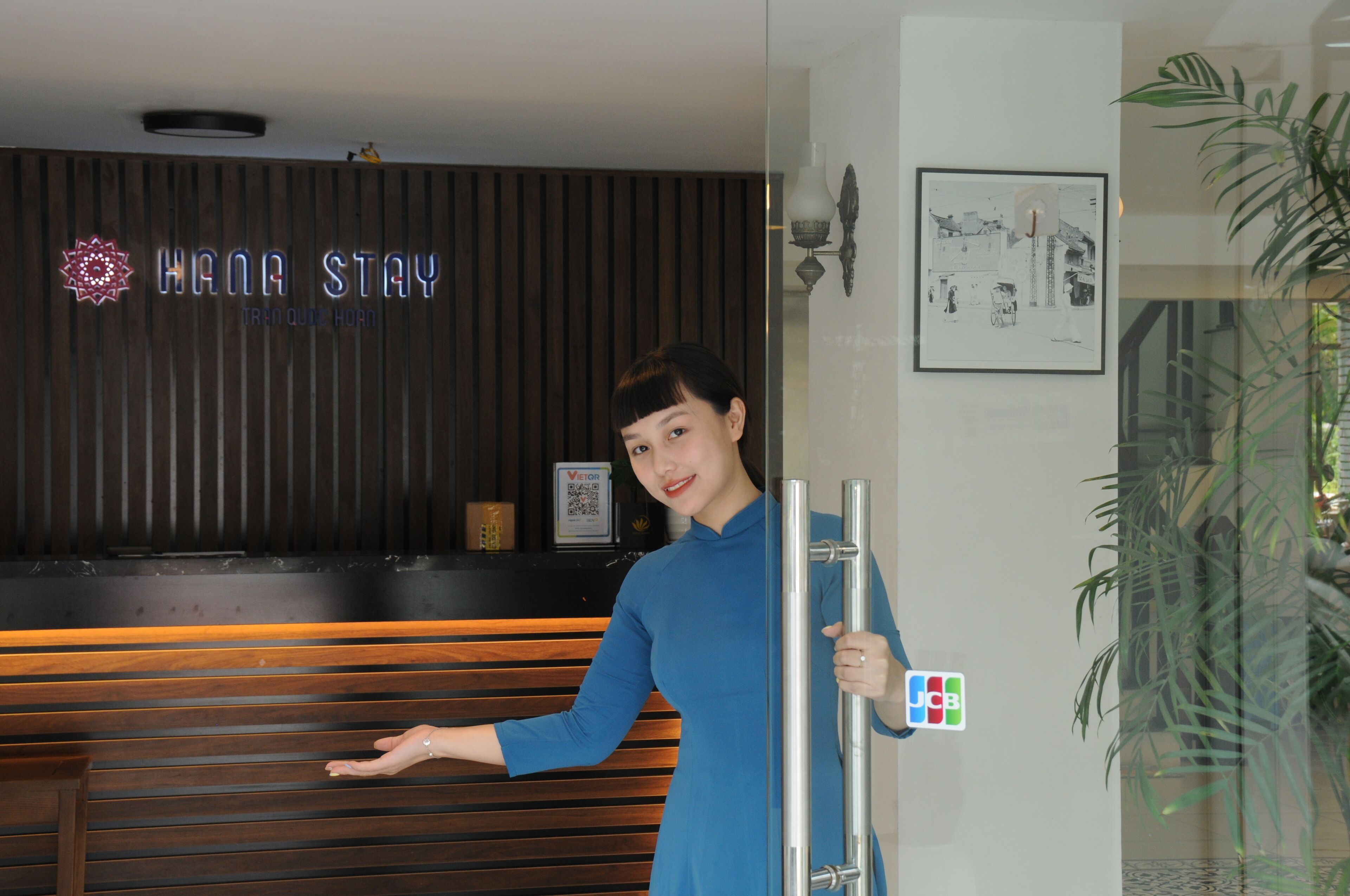 A reception area with a wooden reception desk, a neon sign reading "HANA STAY," and a framed architectural drawing on the wall.