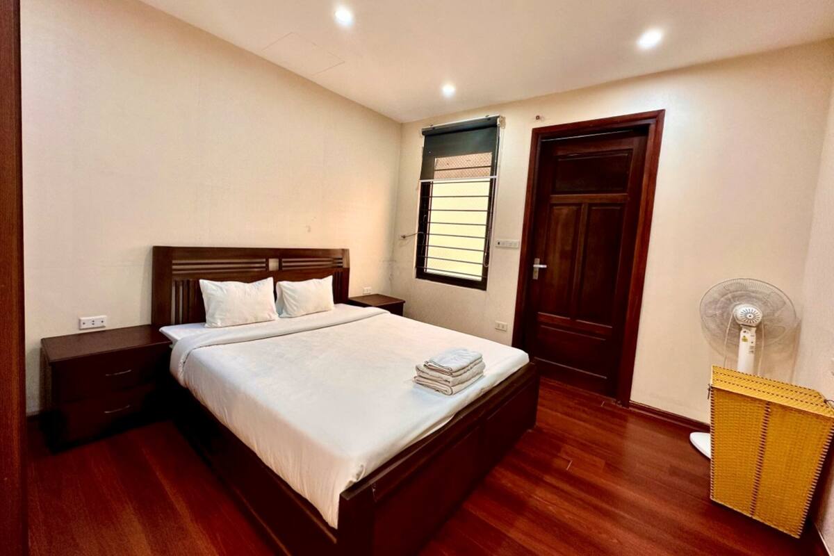 A bedroom with a wooden bed, a nightstand, a window with blinds, a door, and a standing fan.