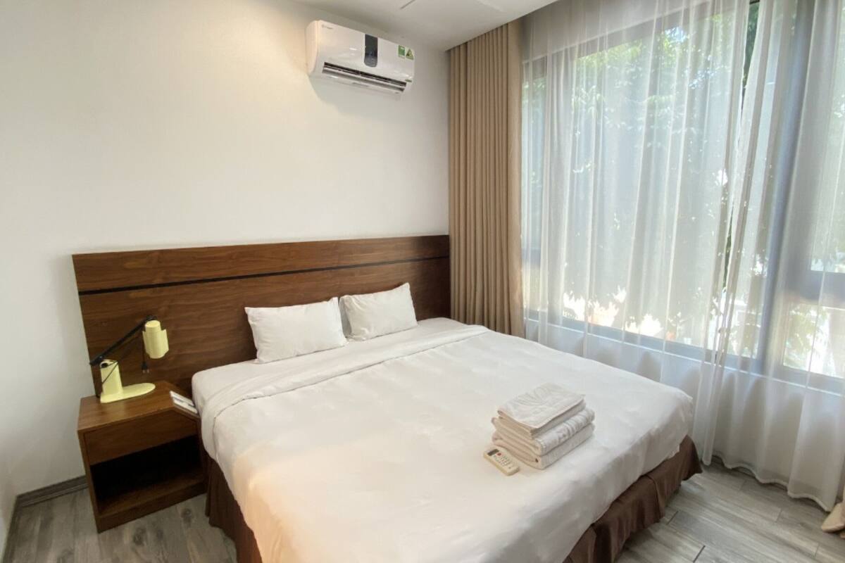 A hotel room with a large bed, a bedside table with a lamp, an air conditioner, and a window with sheer curtains.
