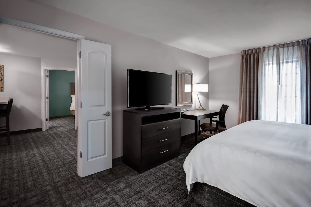 A hotel room with a bed, a desk with a chair, a television, a wardrobe, and a door leading to another room.