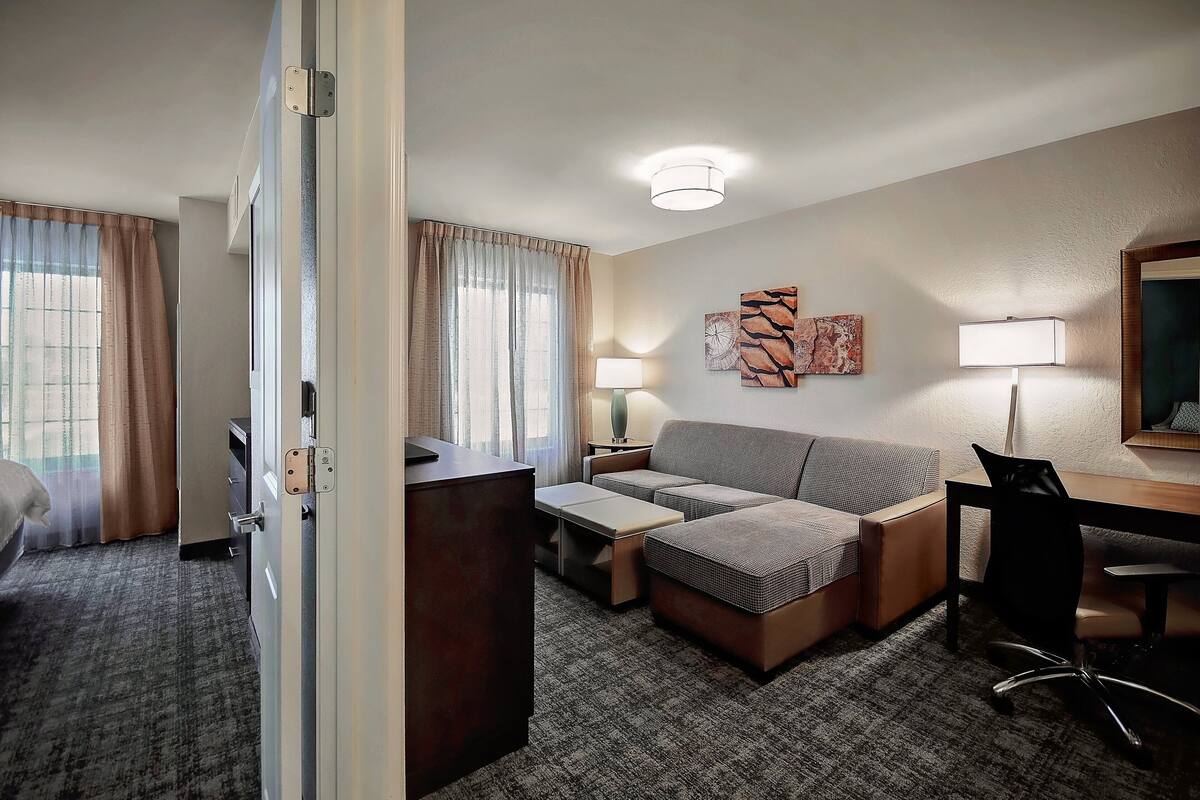 A hotel room with a sofa, a desk, a chair, a lamp, and a painting on the wall.