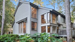 A modern two-story house with large windows and a deck, surrounded by trees and a well-maintained garden. - Island Song Villa - Newly Renovated, Pool, Tennis & Pickleball, Prime Location (Hilton Head Island,)