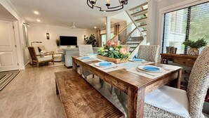 A dining area with a wooden table set for two, a bench, wicker chairs, a chandelier, a staircase, and a TV in the background. - Island Song Villa - Newly Renovated, Pool, Tennis & Pickleball, Prime Location (Hilton Head Island,)
