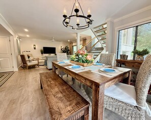 A dining area with a wooden table set for four, a chandelier, wicker chairs, and a staircase in the background.