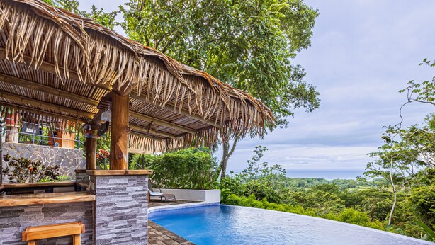 Un área junto a la piscina con un muro de piedra, un banco de madera y una estructura cubierta de paja.