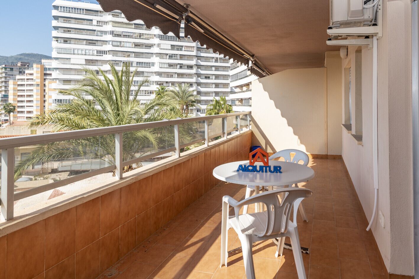 A balcony with a white table and chairs, a logo, and a view of a white building with balconies.