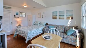 A cozy room with a bed, a sofa, a table, and a chair. There are wall decorations, a clock, and a window with blinds.