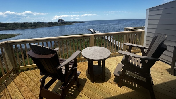 Um deck de madeira com duas cadeiras Adirondack e uma pequena mesa redonda, com vista para um corpo d'água e uma linha de costa distante.