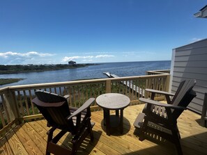 A wooden deck with two Adirondack chairs and a small round table, overlooking a body of water with a distant shoreline. - Shell Shack at The Sea Whisper Condos! (HATTERAS)