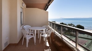 Un balcón con mesa y sillas, con vistas a una playa y una cancha de tenis.