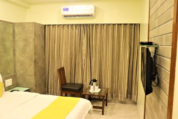 A hotel room with a bed, a chair, a small table with a coffee pot, and a TV mounted on the wall.
