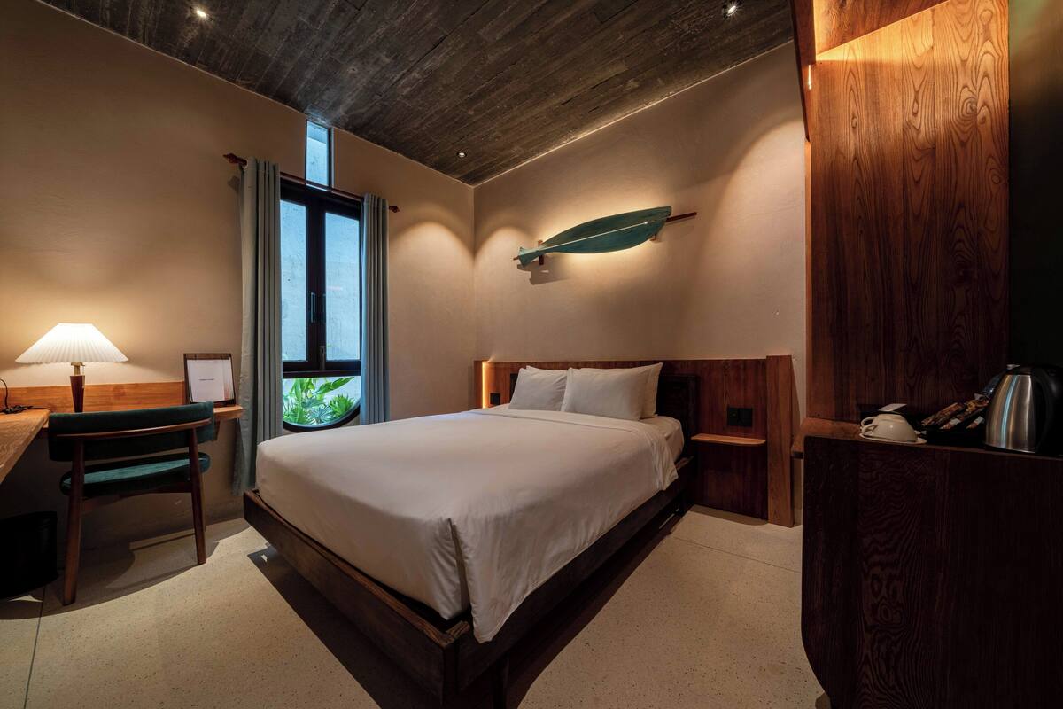 A hotel room with a wooden bed, a desk with a lamp, a window with curtains, and a wall-mounted shelf with a decorative item.