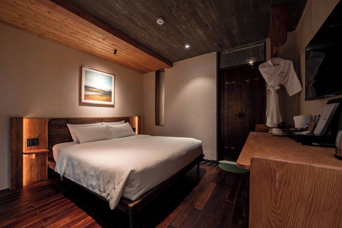 A bedroom with a wooden bed, a bedside table, a robe hanging on the wall, and a wooden floor.